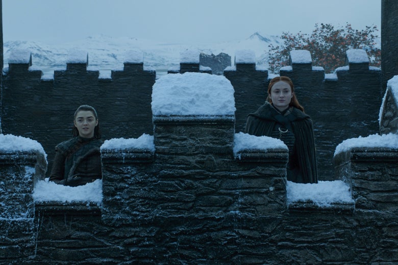 Maisie Williams and Sophie Turner stand behind a snow-covered wall.